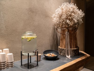 Glass dispenser with lemon water on stand beside paper cups, bowl of nuts, and dried flowers on wooden table