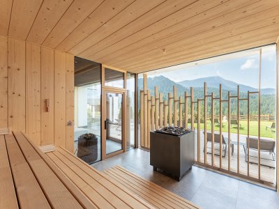 Sala sauna con panche di legno a sinistra e grande finestra con vista su montagne e sedie a sdraio