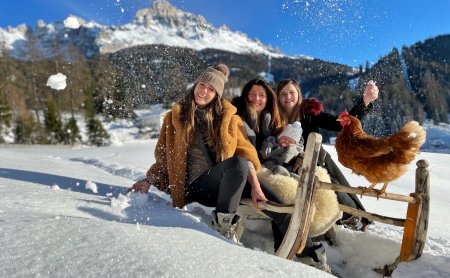 Drei Frauen im Schnee mit Schlitten und Huhn vor Bergkulisse