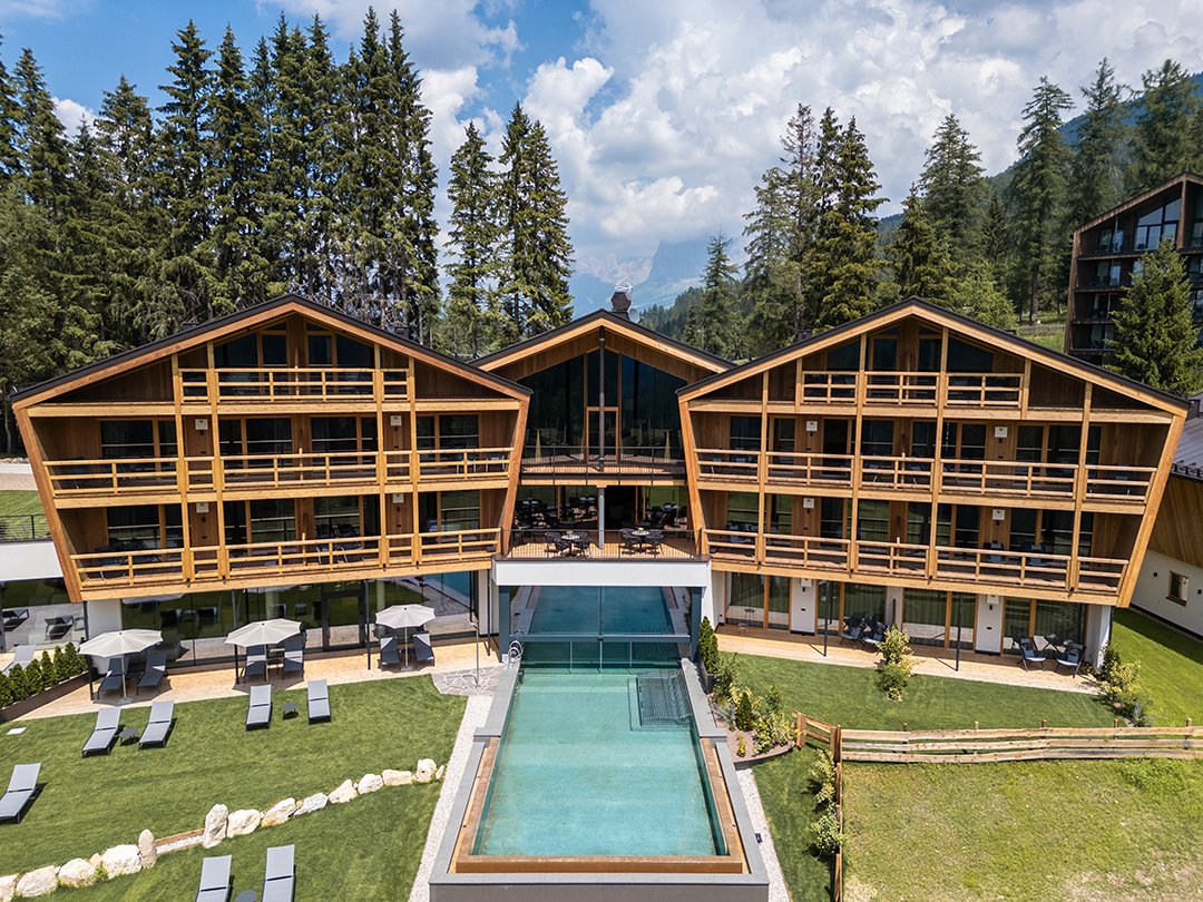 Edifici in legno con balconi intorno a una piscina centrale, circondati da pini e montagne