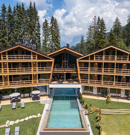 Edifici in legno con balconi intorno a una piscina centrale, circondati da pini e montagne
