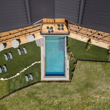 Vista aerea di una piscina accanto a un edificio in legno con lettini, ombrelloni e prati