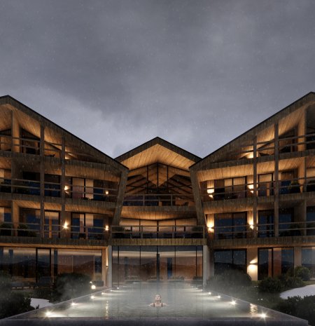 Exterior view of the Family Nature Resort Ortnerhof at dusk. The modern, wood-clad main building is composed of several gable-shaped structures with warmly lit balconies and large glass façades. In the foreground, a softly illuminated outdoor pool emits a light mist. A dramatic, cloud-filled sky stretches above the resort.