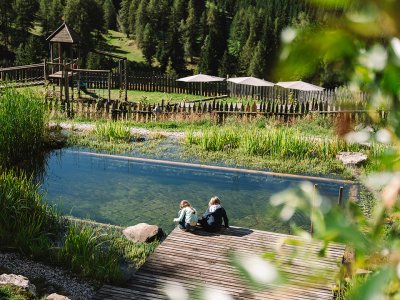Two children sit on a wooden dock by a pond with a playground and trees behind