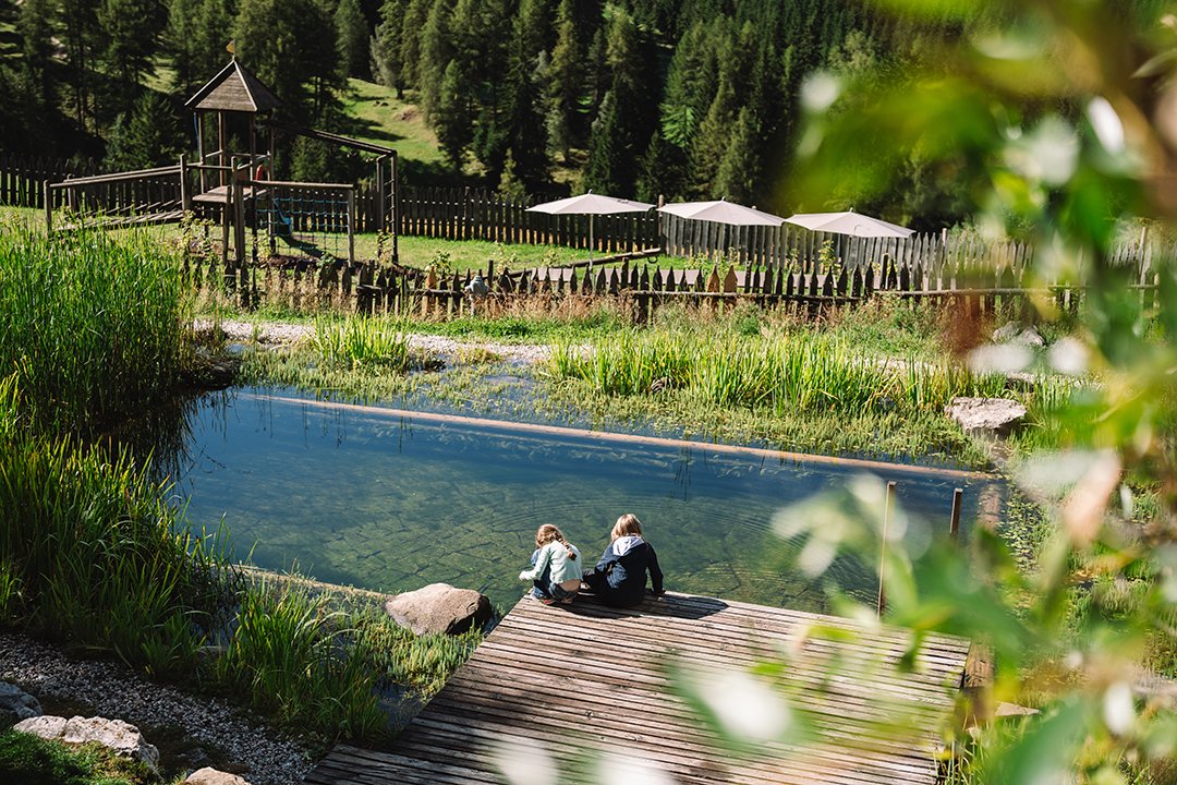 Due bambini seduti su un molo di legno accanto a uno stagno con un parco giochi e alberi sullo sfondo