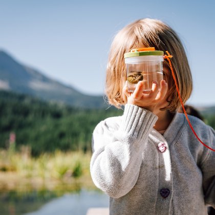 Kind hält Becherlupe mit Frosch am Teich
