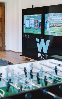 Tischfußball im Vordergrund, im Hintergrund zwei Bildschirme mit Fußballspiel und Text 'Great Save!'