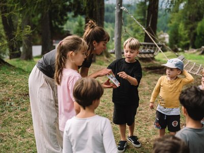 Frau zeigt Kindern im Freien ein Bild, umgeben von Bäumen und Spielplatz