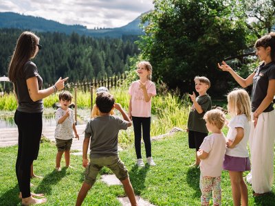 Zwei Erwachsene und mehrere Kinder stehen im Kreis auf einer Wiese vor Wald und Bergen und zeigen mit den Fingern.