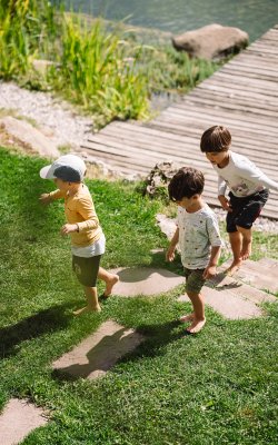 Three children barefoot walking on a grassy path near a wooden dock by the water