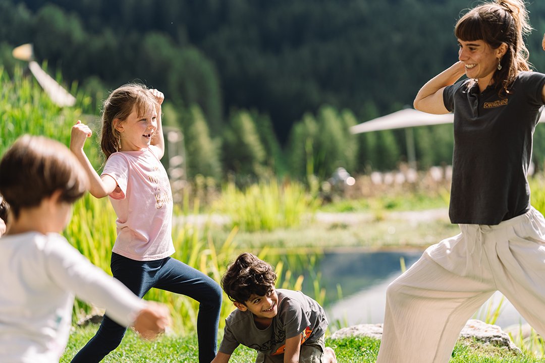 Frau und Kinder machen draußen Yoga-Übungen auf einer Wiese vor einem Teich