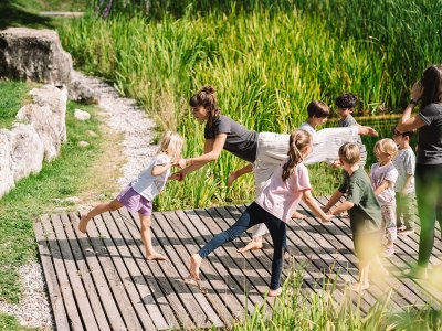 Woman doing yoga pose on wooden dock holding children's hands, other kids and woman watching