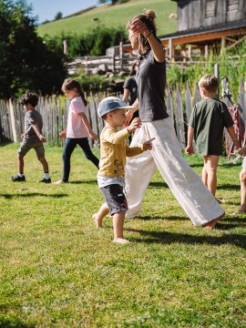 Kinder und eine Frau tanzen barfuß auf einer Wiese vor einem Holzzaun und Bauernhof