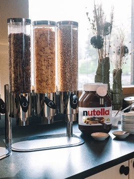 Frühstückstheke mit Getreidespendern, großem Nutella-Glas und Schalen vor Fenster mit Trockenblumen