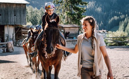 Kind reitet braunes Pferd, begleitet von lächelnder Reitlehrerin