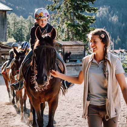 Kind reitet braunes Pferd, begleitet von lächelnder Reitlehrerin
