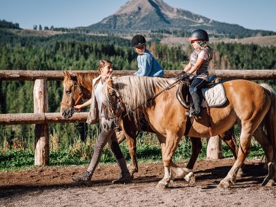 Zwei Kinder reiten Pferde mit Reitlehrerin vor grünem Berghintergrund