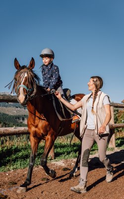 Kind auf braunem Pferd mit Reitlehrerin am sonnigen Reitweg