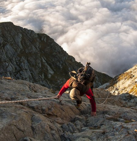 Kletterer zieht sich mit Seil über Felswand hoch, Wolkenmeer im Hintergrund