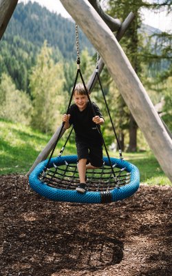 Kind spielt auf einer blauen Nestschaukel im Wald mit Bergen im Hintergrund