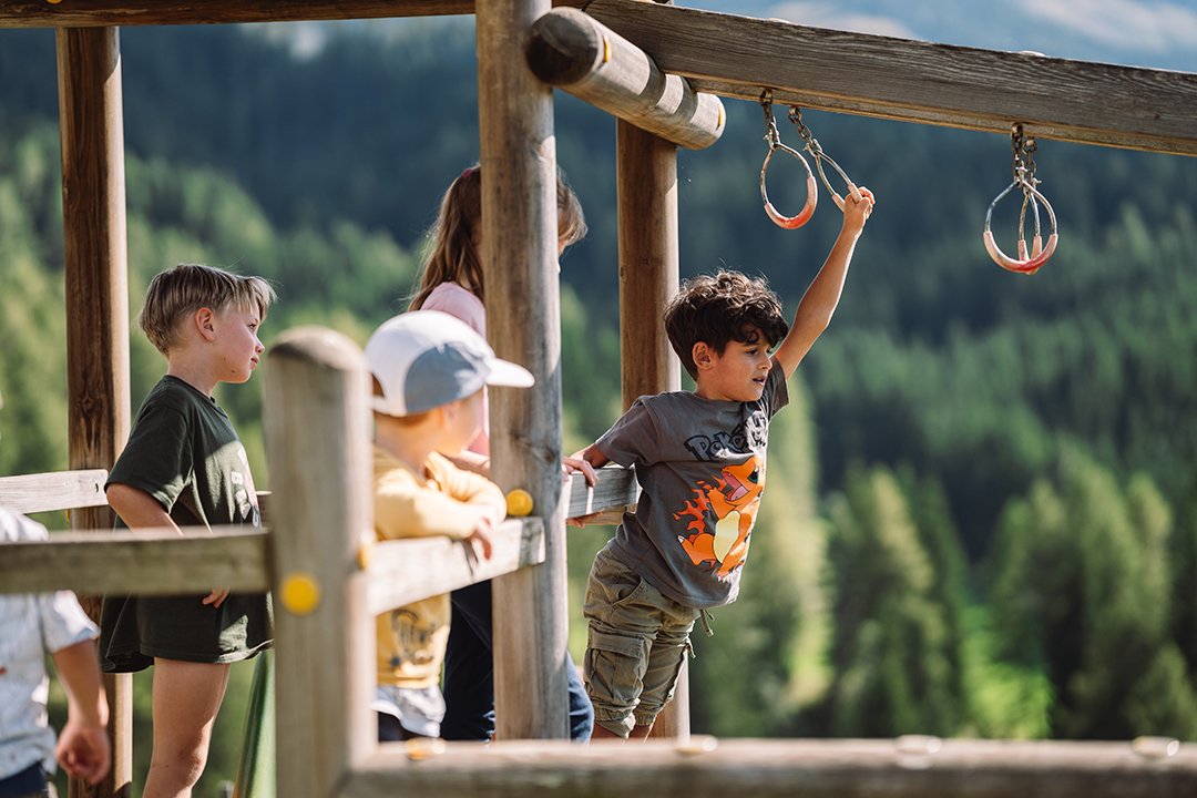 Children playing on a wooden playground with rings against a forested mountain background