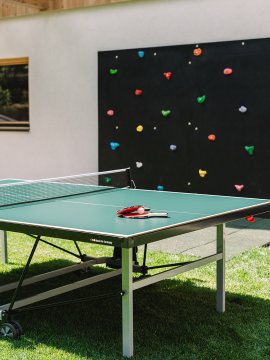 Tischtennisplatte mit Schlägern und Ball auf grünem Rasen vor Kletterwand mit bunten Griffen