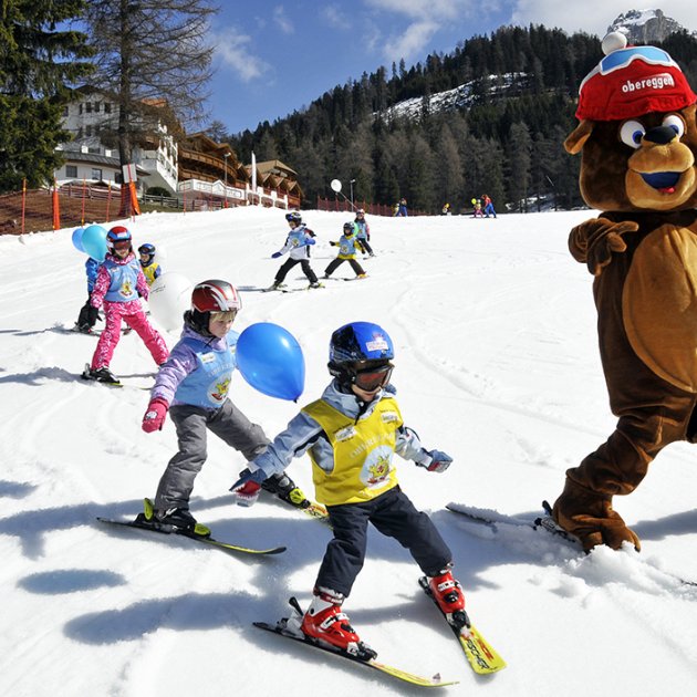 Orso mascotte scivola sulla neve con bambini che imparano a sciare e palloncini colorati.