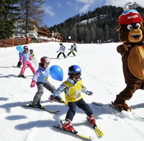 Orso mascotte scivola sulla neve con bambini che imparano a sciare e palloncini colorati.