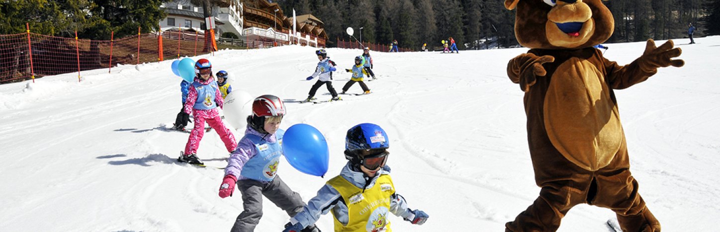 Maskottchen-Bär fährt auf Schnee mit Kindern, die Skifahren lernen, und bunten Luftballons.
