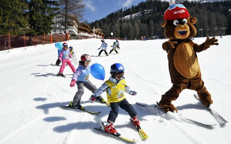 Maskottchen-Bär fährt auf Schnee mit Kindern, die Skifahren lernen, und bunten Luftballons.