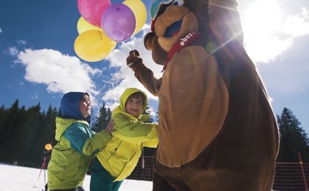 Maskottchen-Bär mit gelben und violetten Luftballons spielt mit Kindern im Schnee.