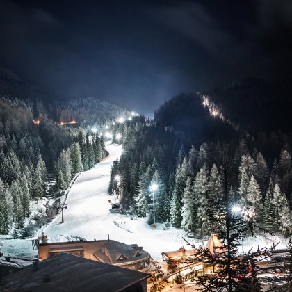 Beleuchtete Skipiste bei Nacht, umgeben von Tannen und verschneiten Bergen.