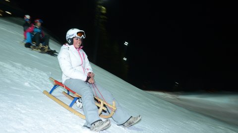 Frau rodelnd bei Nacht auf verschneiter Piste mit weiteren Personen dahinter.