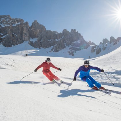 Zwei Skifahrer in roten und blauen Anzügen fahren eine verschneite Piste mit felsigen Bergen und strahlender Sonne hinunter.