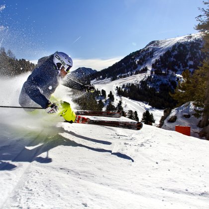 Skifahrer mit blauer Jacke und gelber Hose fährt schnell eine verschneite Piste hinunter und wirbelt Schnee auf.
