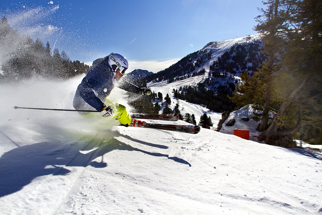 Skifahrer mit blauer Jacke und gelber Hose fährt schnell eine verschneite Piste hinunter und wirbelt Schnee auf.