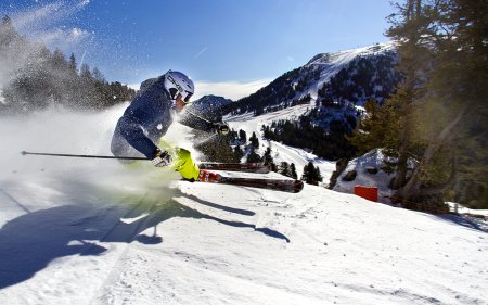 Skifahrer mit blauer Jacke und gelber Hose fährt schnell eine verschneite Piste hinunter und wirbelt Schnee auf.