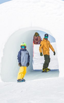 Schneetunnel mit Kind und Erwachsenem in Skihelm und Winterkleidung.