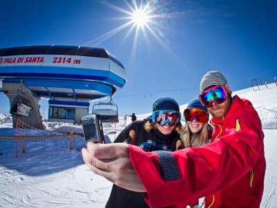 Drei Personen mit Skijacken und Sonnenbrillen machen ein Selfie auf einer verschneiten Piste neben einem Sessellift.