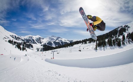 Snowboarder mit gelber Jacke und schwarzer Hose macht einen hohen Sprung in einem Snowpark mit Bergen im Hintergrund.