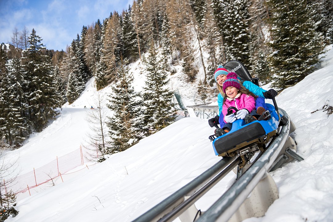 Mutter und Kind fahren lachend auf rotem Sommerrodel im Schnee.