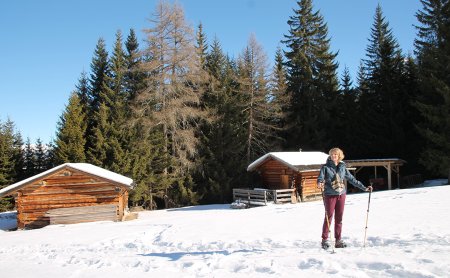 Frau mit Wanderstöcken steht im Schnee vor verschneiten Holzhütten.