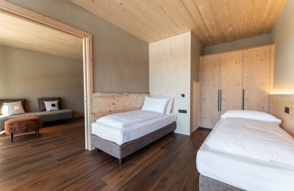 Two single beds in a room with wooden walls and floor, adjacent to a living room with a sofa